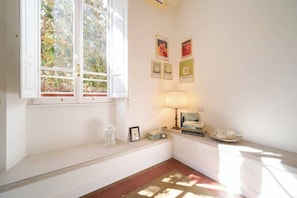 A room with a window, a lamp, a shelf with books, and framed pictures on the wall.