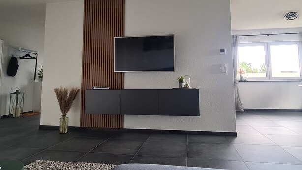 A modern living room with a flat-screen TV mounted on a wall, a dark wooden accent wall, a black entertainment unit below, and a large window providing natural light. - M&M AusZeit in Schwabenländle in Winterlingen (Winterlingen)