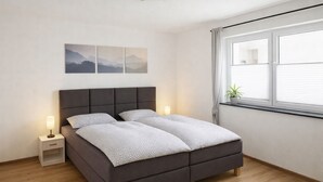 A modern bedroom with a dark brown bed, a white bedside table, and a window with blinds. - M&M AusZeit in Schwabenländle in Winterlingen (Winterlingen)