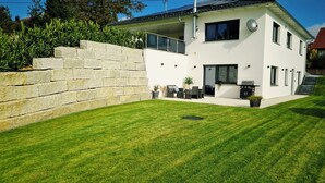 A modern two-story house with a stone retaining wall, a well-maintained lawn, and a patio area with outdoor furniture. - M&M AusZeit in Schwabenländle in Winterlingen (Winterlingen)