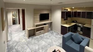 A modern living room with a flat-screen TV, a coffee table, and a sofa. There is a kitchen area with cabinets and a countertop. - Villa park (Al Khobar)