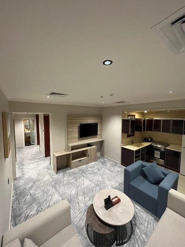 A modern living room with a flat-screen TV, a coffee table, and a sofa. There is a kitchen area with cabinets and a countertop. - Villa park (Al Khobar)