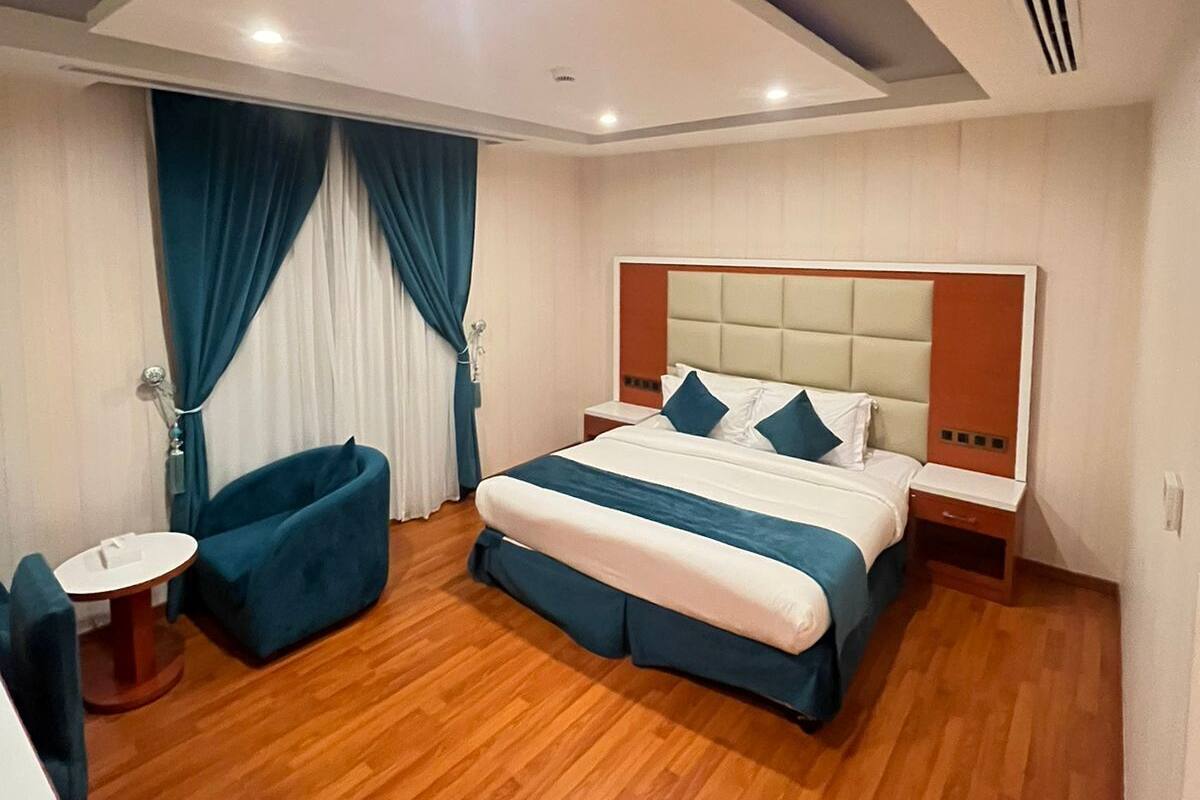 Una habitación de hotel moderna con una cama grande, una mesita redonda pequeña, un sillón azul y una rejilla de ventilación en el techo.