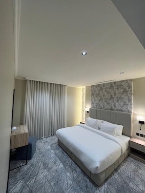 Quarto de hotel moderno com uma cama grande, uma mesa de cabeceira, uma luminária e uma janela com cortinas.