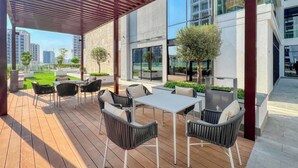 Un'area patio esterna con pavimento in legno, sedie in vimini nero e tavoli bianchi.