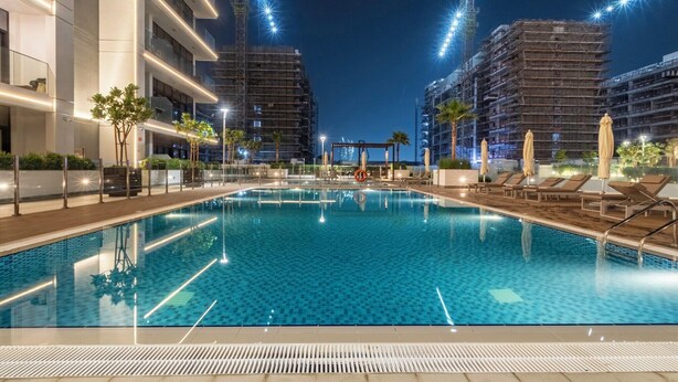 Una piscina con lettini e ombrelloni, circondata da edifici moderni e gru da cantiere.