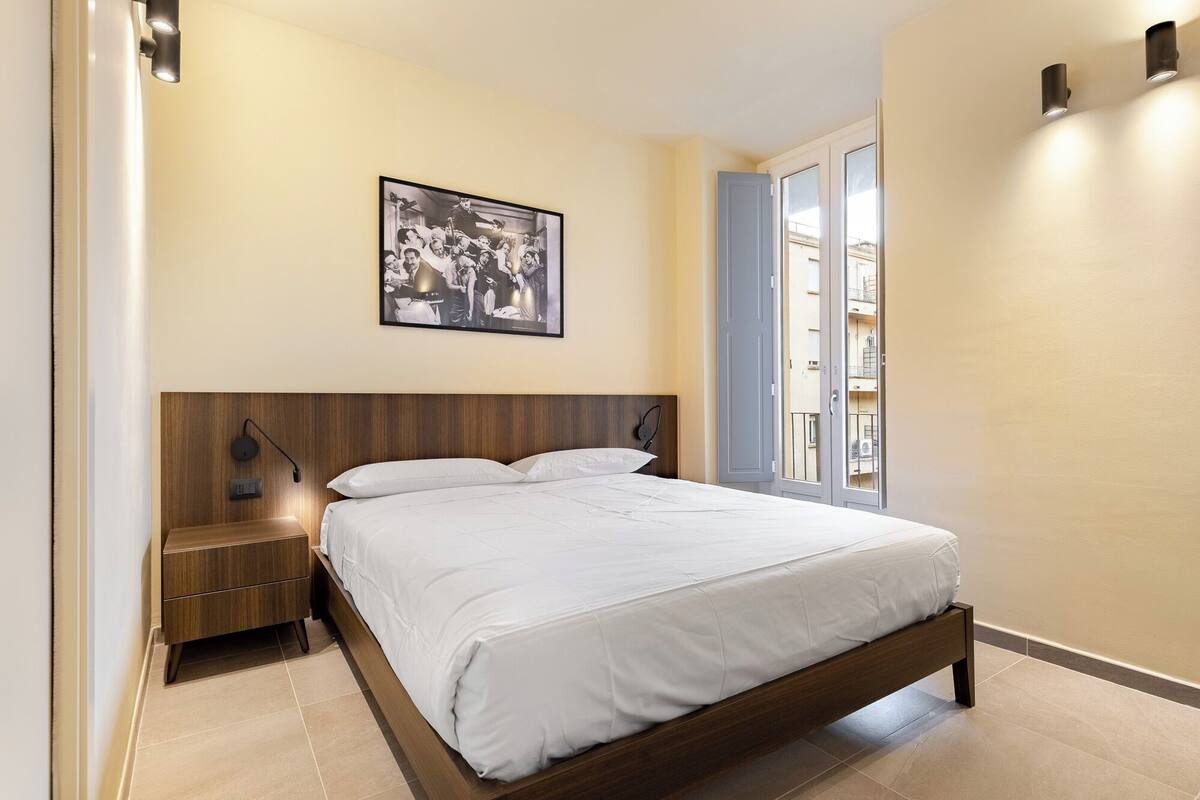 A bedroom with a wooden bed, a nightstand, a wall-mounted lamp, and a framed picture above the bed.