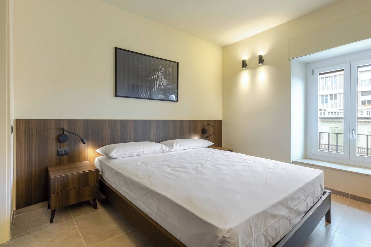 A neatly arranged bedroom with a bed, bedside tables, a wall-mounted artwork, and a window with a view of buildings.