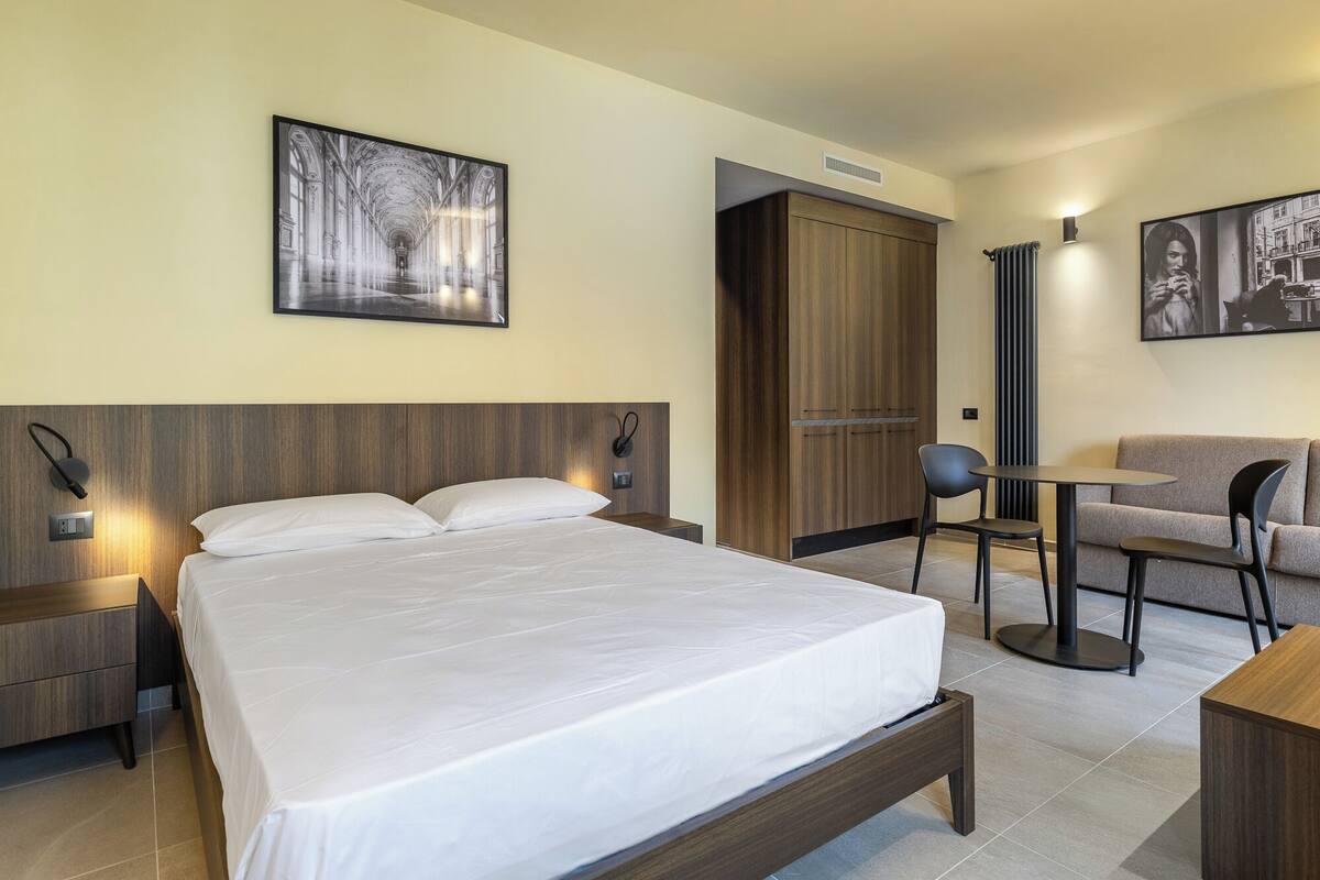A modern hotel room with a large bed, a wooden wardrobe, a seating area with a table and chairs, and artwork on the walls.