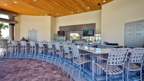 A covered outdoor bar area with metal chairs and a counter, a kitchen in the background, and a door.