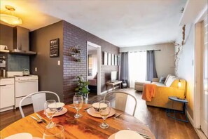 A dining area with a wooden table set for four, a kitchen with modern appliances, a living area with a sofa and a lamp, and a brick accent wall. - King Bed in MidtownWalk to Alessi Bakery & Midtown (Tampa)