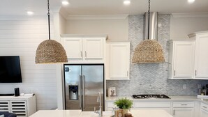 A modern kitchen with a central island, white cabinetry, stainless steel appliances, and wicker pendant lighting. - Seas the Day 30A - 6 Seater Golf Cart, 2 Pools, Beach Chairs, Wi-Fi & AC (Panama City Beach)