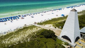 A beach with white sand, blue umbrellas, and a tall white building with a pointed roof. - Seas the Day 30A - 6 Seater Golf Cart, 2 Pools, Beach Chairs, Wi-Fi & AC (Panama City Beach)