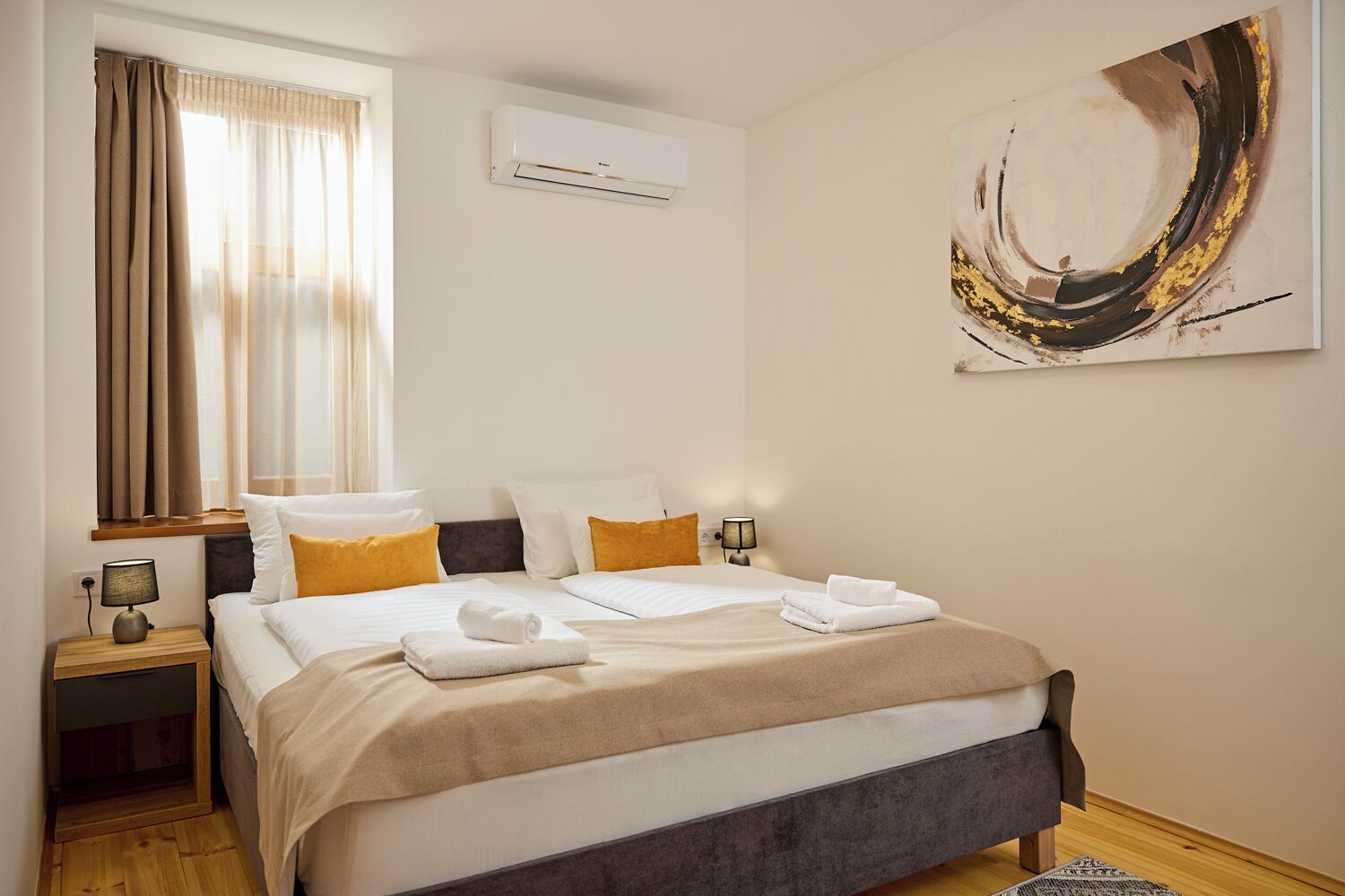 A hotel room with a bed, a window with curtains, an air conditioner, a lamp, and a painting on the wall.