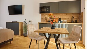 A modern kitchen with dark cabinets, a microwave, and a sink. There is a dining table with two chairs and a vase with flowers. A TV is mounted on the wall, and there is a bed in the corner. - Villa Olea (Tihany)