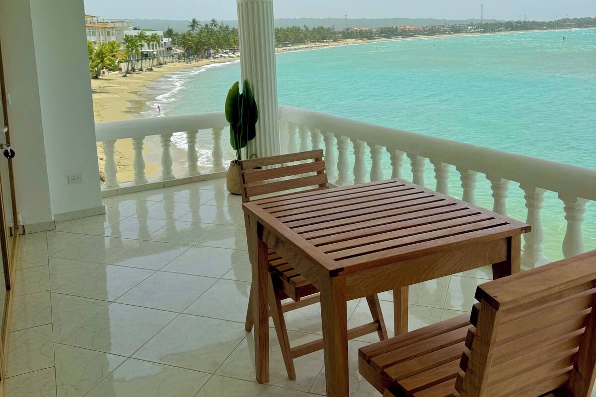 A balcony with a wooden table and chairs, overlooking a beach with clear turquoise water and a distant shoreline.