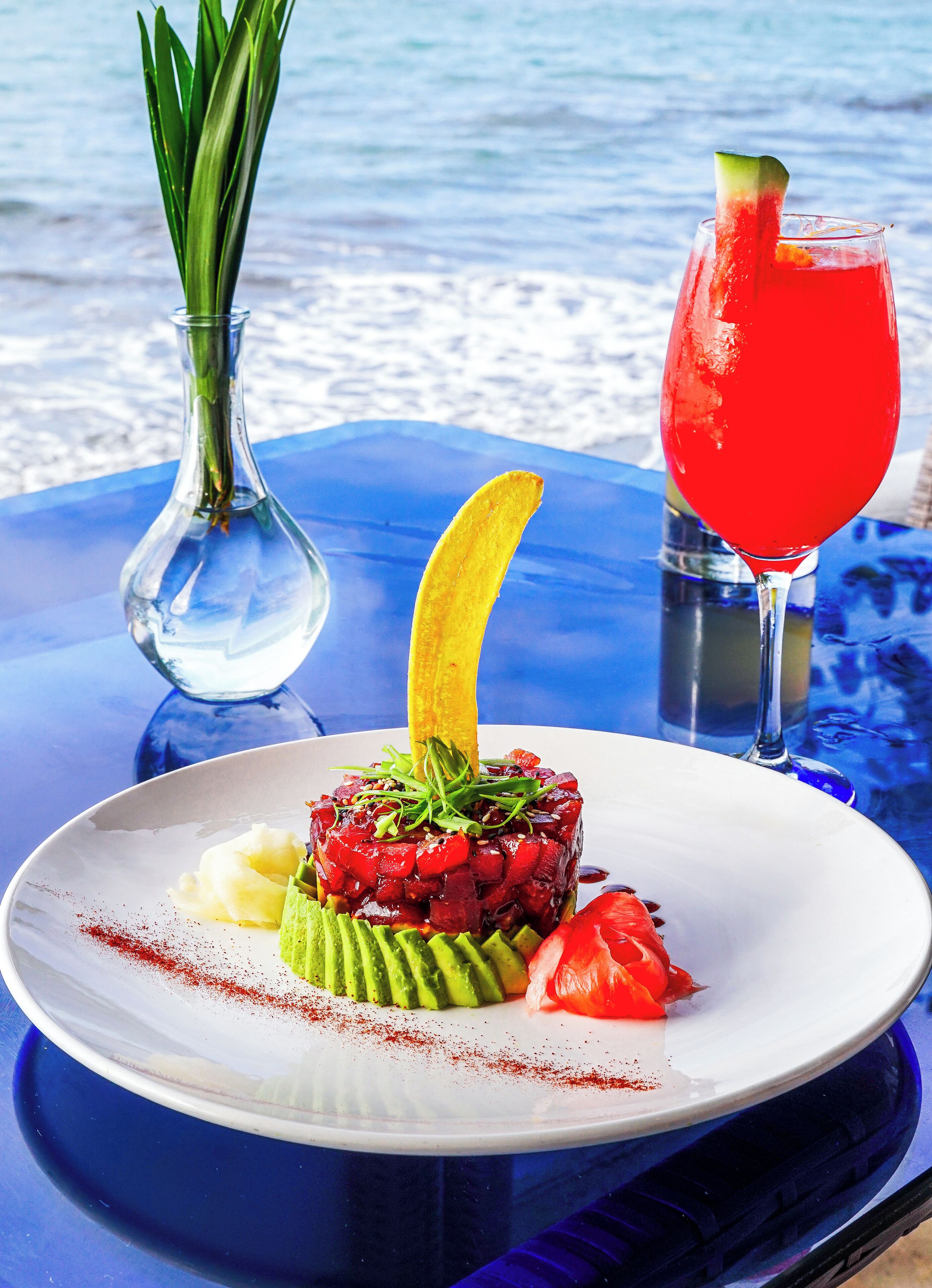 A seafood dish on a white plate with avocado, tomato, and a decorative yellow element, accompanied by a red drink in a glass.