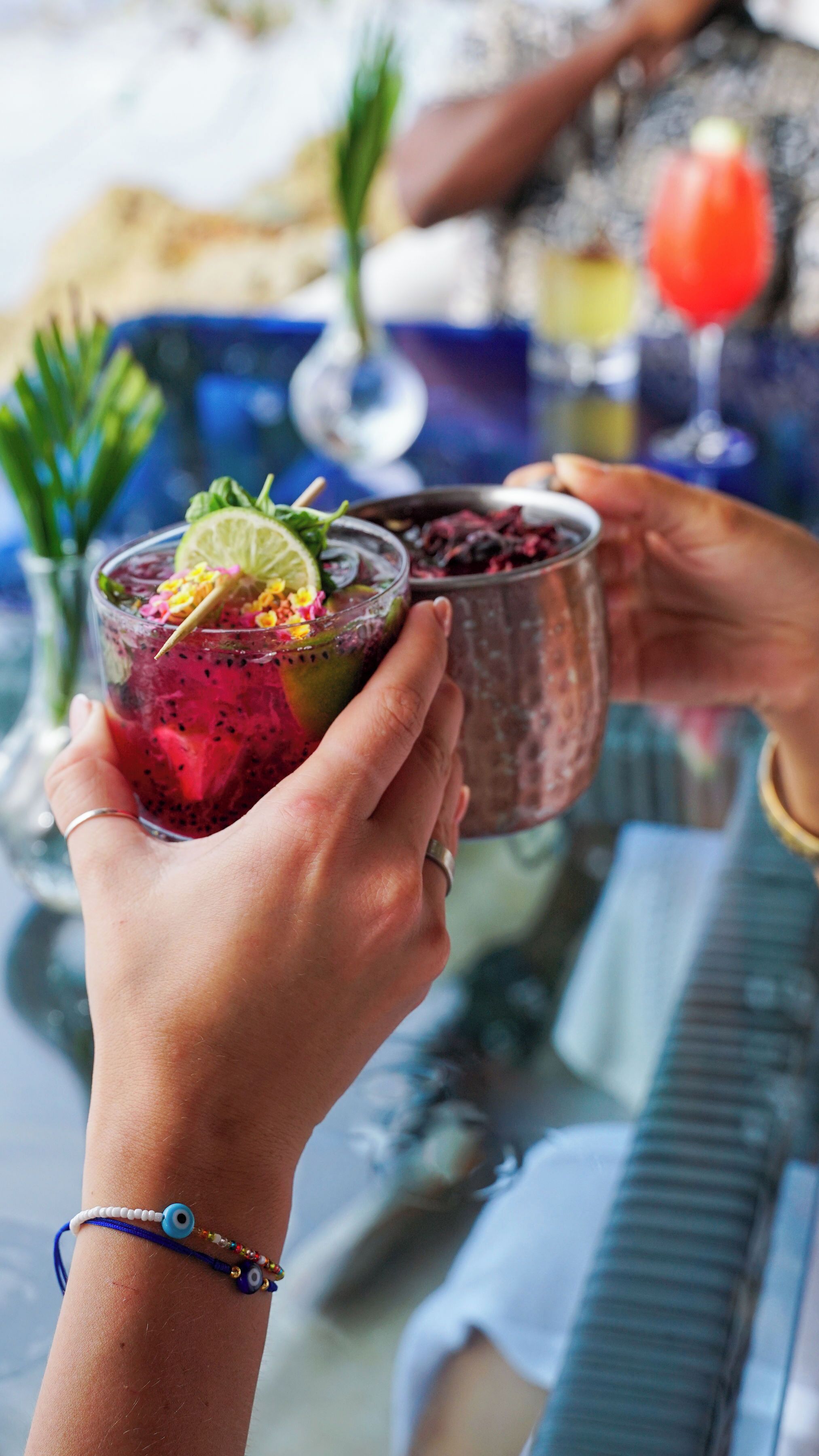 A person holding a glass with a colorful drink, another person in the background holding a different drink.
