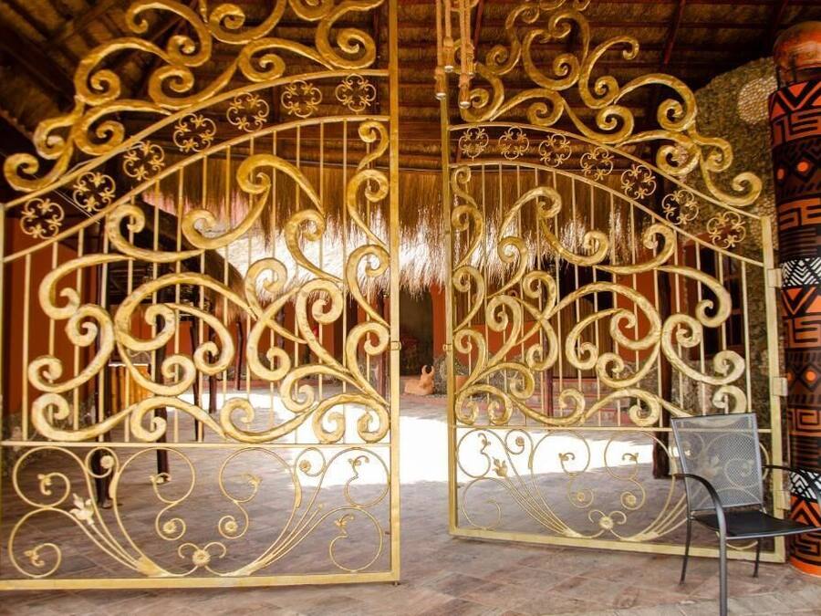 Une porte en métal décorative, ornée de motifs complexes, qui donne accès à un espace intérieur doté d’un sol à motifs et d’une chaise.
