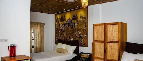 Habitación de hotel con dos camas, un armario de madera, una mesita, una silla, un hervidor rojo y una pared con un mural decorativo.
