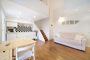 A modern kitchen with a checkered backsplash, a dining area with a table and chairs, a white sofa, and a staircase leading to an upper level.