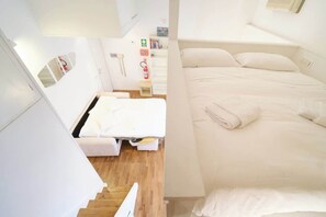 Une chambre d’hôtel avec un lit, une petite table de chevet, une étagère fixée au mur avec des livres et un extincteur.