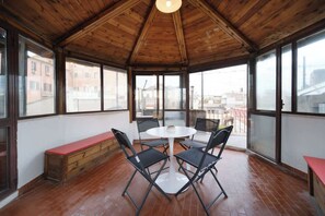A room with a wooden ceiling, large windows, a round table with chairs, a bench, and a red table.