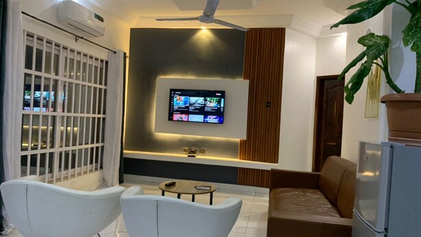 A modern living room with a flat-screen TV, white chairs, a brown sofa, and a refrigerator.