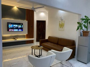 Een moderne woonkamer met een bruine bank, witte fauteuils, een klein rond tafeltje, een flatscreen televisie aan de muur en een potplant.