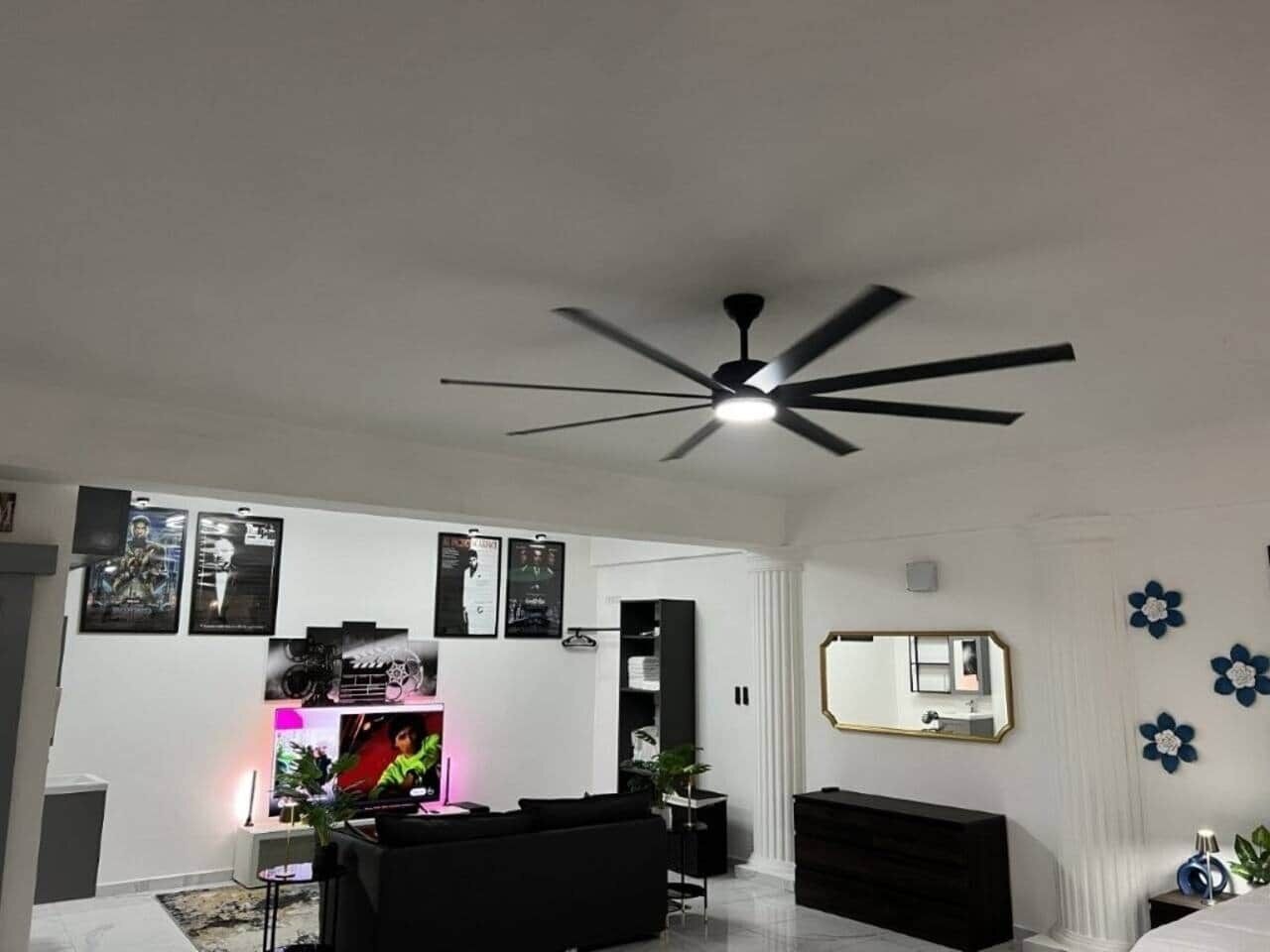 Un salon moderne avec un ventilateur de plafond, un espace dédié aux jeux vidéo, un canapé noir et un grand miroir.