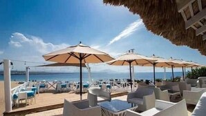 Un espace extérieur aménagé avec des meubles en osier blanc et des parasols beige, offrant une vue sur une étendue d’eau.