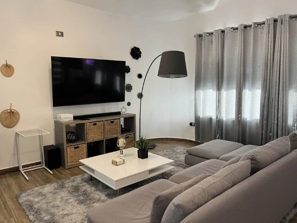 Un soggiorno moderno con una televisione a schermo piatto, un tavolino da caffè bianco, un divano grigio, una lampada da terra nera e una tenda grigia.