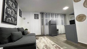 A modern living room with a grey sofa, a small rug, a refrigerator, and a dining area with a table and chairs. - The Driftwood Hideaway (Sosúa)