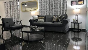 Un salon moderne avec un canapé en cuir noir, un fauteuil noir et une table basse ronde. On y trouve deux tables d’appoint avec des lampes, un grand miroir et des rideaux à motifs.