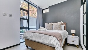 A modern bedroom with a large bed, a bedside table, and a window with a view of trees.