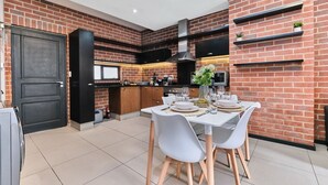 A modern kitchen with brick walls, a dining area with white chairs and a table set for four, and built-in shelves with plants.