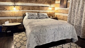 Un dormitorio acogedor con pared de troncos de madera, una mesa redonda, una cama con ropa de cama floral y una mesita de noche con lámpara.