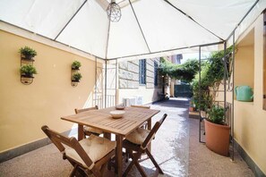 Un patio cubierto con una mesa y sillas de madera, plantas en macetas y vista a la calle.