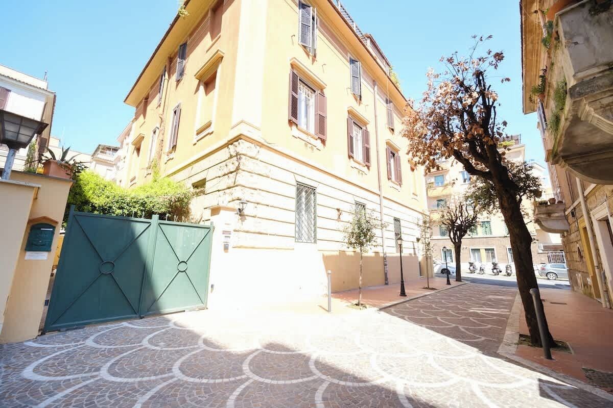 Un edificio con un patio empedrado, una verja verde y un árbol de follaje escaso.