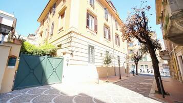 Un edificio con un patio empedrado, una verja verde y un árbol de follaje escaso.