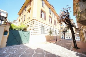 Un edificio con un patio empedrado, una verja verde y un árbol de follaje escaso.