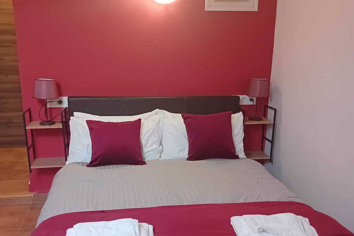 Ein Bett mit roter und weißer Bettdecke, zwei Kissen und zwei gefaltete Handtücher. Zwei Nachttischlampen und ein gerahmtes Bild sind an der Wand zu sehen.
