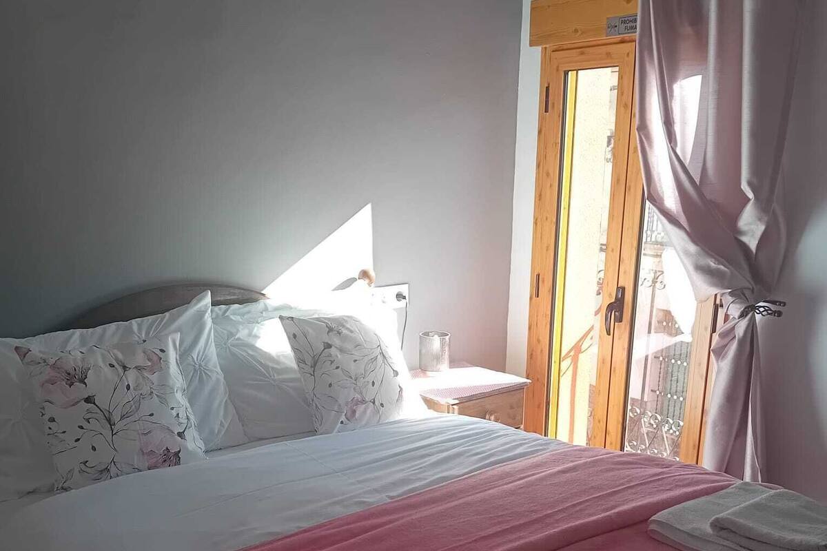 Ein ordentlich bezogenes Bett mit roter Decke und weißen Kissen, ein kleiner Nachttisch mit Lampe sowie ein Fenster mit Vorhängen.