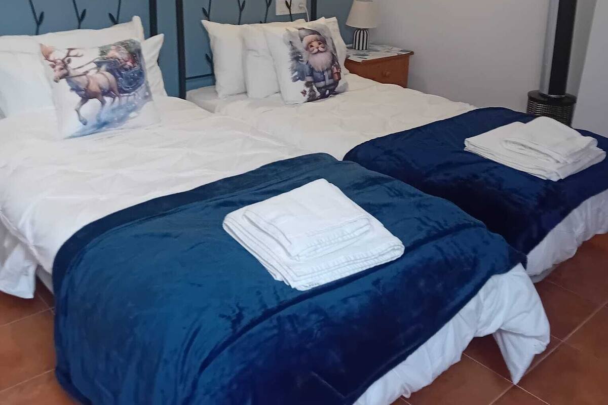 Ein Zimmer mit zwei Betten, jeweils mit blauer Bettdecke und weißer Bettwäsche. Es gibt einen Nachttisch mit einer Lampe, ein gerahmtes Bild an der Wand und eine Heizung.