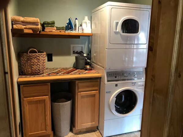 A compact laundry area with a stacked washer and dryer, a shelf with folded towels, a basket, and a trash can.