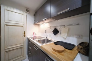 Una cucina compatta con una porta bianca, mobili scuri, un lavello in acciaio inox, un piano di lavoro in legno e una rastrelliera pensile per pentole.