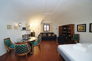 A hotel room with a bed, a dining table with chairs, a sofa, a bookshelf, and a view of the outside through a window.