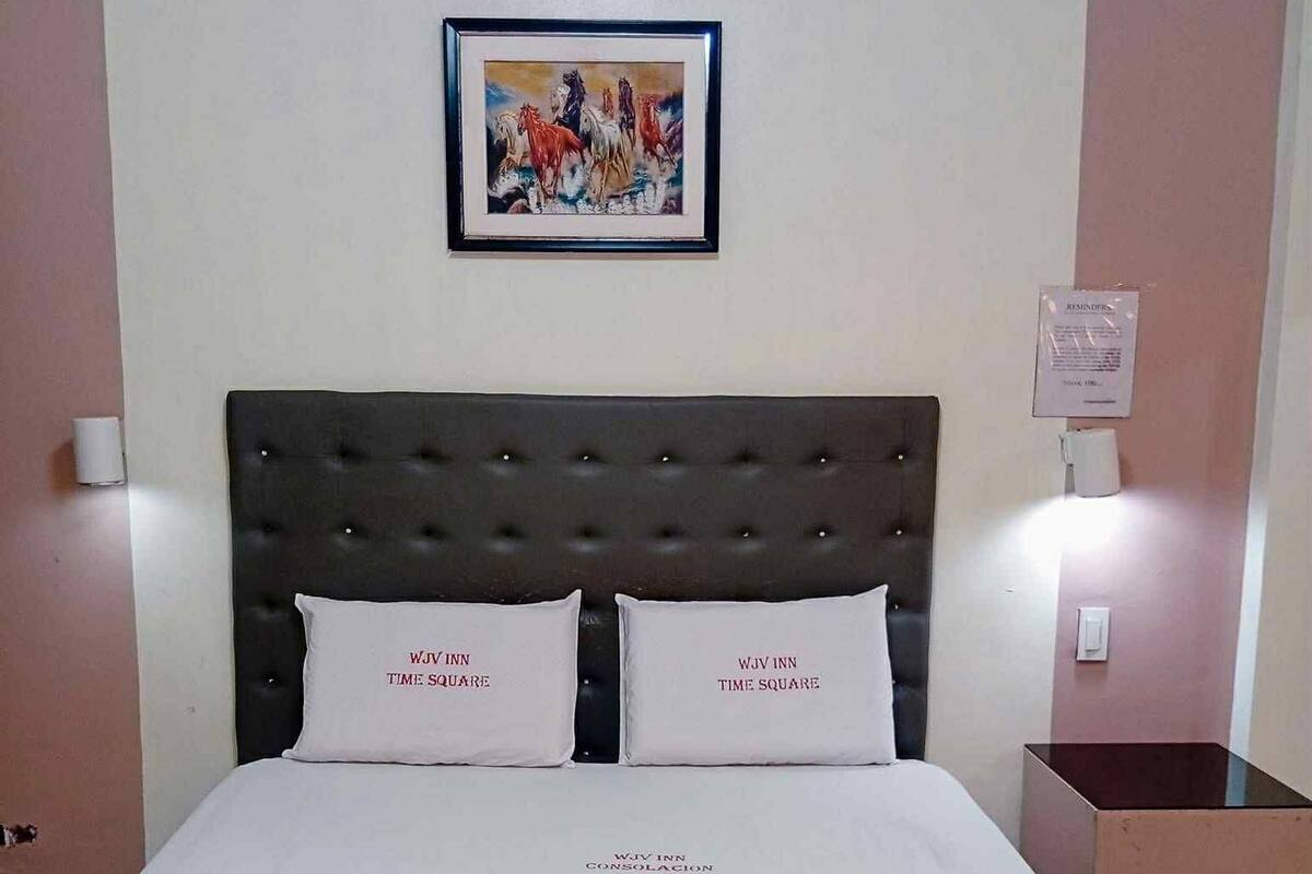 A hotel room with a large bed, two pillows labeled "WJW INN TIME SQUARE", a framed picture on the wall, and a bedside table with a lamp.