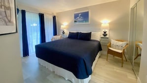 Une chambre avec un grand lit, deux tables de chevet avec des lampes, un fauteuil en osier, un tableau encadré au mur et une fenêtre avec des rideaux.