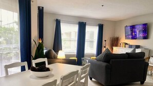 A modern living room with a white dining table, a dark grey sofa, and a flat-screen TV mounted on the wall. - SeaWorld Orlando | 2BR | Minutes to the Parks (Orange County)